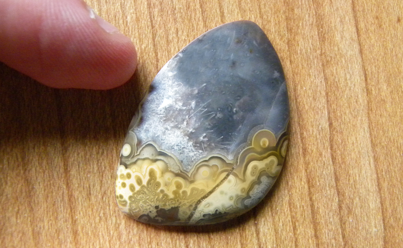 Yellow tube and eye patterns below a dark blue-gray sky and white needles in a freeform cabochon of Mexican Crazy Lace Agate.