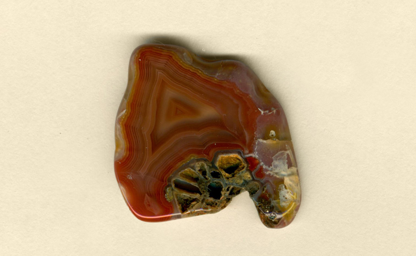 A freeform Coyamito Agate from Mexico, with an orange and red triangular fortification pattern dominating it, resting on the beginnings of an agate pseudomorph after aragonite. 